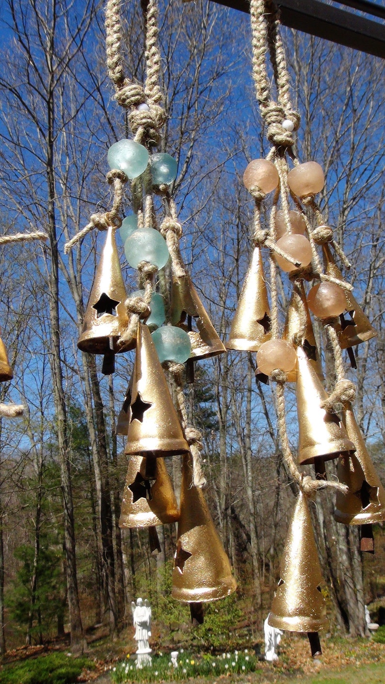 May include: A set of gold-colored bells hanging from a rope with blue and pink glass beads. The bells are cone-shaped with a star cut-out and are hanging in a forest setting.