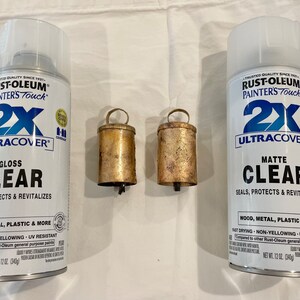 May include: Two cans of Rust-Oleum Painters Touch 2X Ultracover clear spray paint. One can is gloss and the other is matte. There are two small, gold-colored bells in the center of the image.