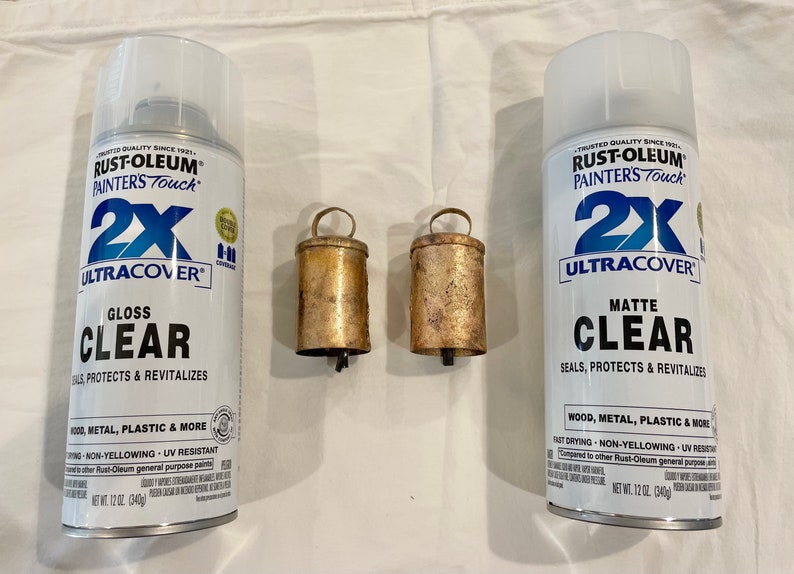May include: Two cans of Rust-Oleum Painters Touch 2X Ultracover clear spray paint. One can is gloss and the other is matte. There are two small, gold-colored bells in the center of the image.
