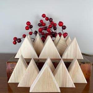 May include: A set of 12 unfinished wooden triangle shapes, arranged in a staggered pattern, resembling a miniature forest. The triangles are made of natural wood and have a smooth, unpainted surface. The triangles are displayed on a brown book with a red berry garland behind them.