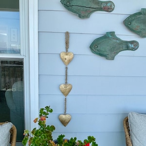 Puede incluir: Una guirnalda de corazones dorados con tres corazones colgando de una cuerda de yute. La guirnalda está colgada en una pared blanca con tres decoraciones de peces verdes. Una planta en maceta con flores rojas está en primer plano.