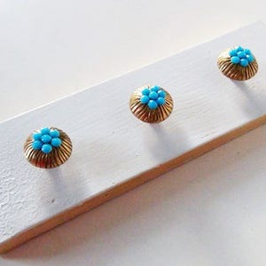 May include: Three gold-colored hooks with blue flower-shaped accents mounted on a white wooden board.