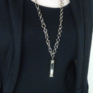 May include: A silver chain necklace with a small, cylindrical pendant. The pendant has a clear glass tube with black specks inside.