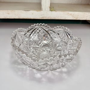 May include: A clear, cut-glass bowl with a scalloped rim. The bowl features a detailed geometric pattern of starbursts, diamonds, and other shapes. The bowl is likely vintage and is sitting on a white surface.