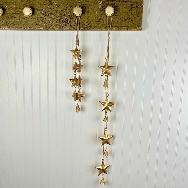 May include: Two gold star and bell wind chimes hanging from a rustic green wooden board. The chimes are made of metal and have a distressed finish.