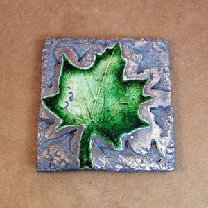 Può includere: Una piastrella quadrata in ceramica con un design a foglia d'acero verde. La piastrella ha uno sfondo argentato testurizzato.