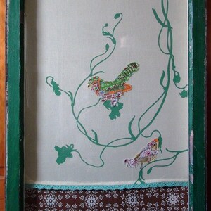 May include: A green wooden frame with a white background and a brown patterned border. The frame has a green and white floral design with two birds made of fabric. The frame has five decorative knobs.