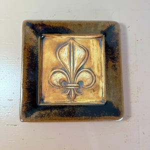 Può includere: Piastrella quadrata in ceramica con un motivo a giglio in rilievo. La piastrella ha un centro dorato con un bordo marrone scuro. Il giglio è un simbolo della regalità francese. La piastrella è probabilmente decorativa.