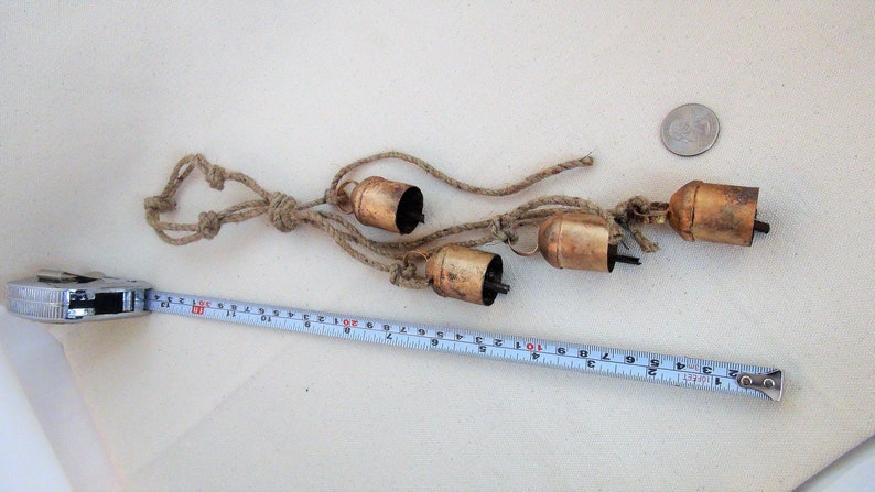 Peut inclure: Un ensemble de quatre petites cloches en laiton d'aspect antique suspendues &agrave; une longueur de ficelle. Les cloches sont li&eacute;es ensemble par des n&oelig;uds et mesurent environ 30 cm de long.