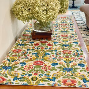 May include: A floral patterned table runner with a cream background, featuring red, blue, and yellow flowers and foliage. A glass vase with dried flowers sits on a wooden stand. The runner is placed on a wooden table.