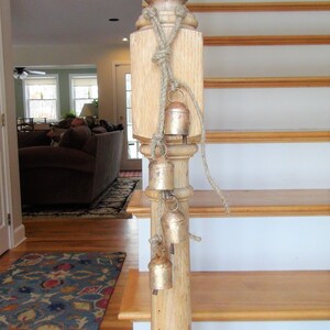 Peut inclure: Une main courante en bois avec une corde et une s&eacute;rie de petites cloches dor&eacute;es qui pendent. La main courante fait partie d'un escalier avec des marches en bois et des contremarches blanches.