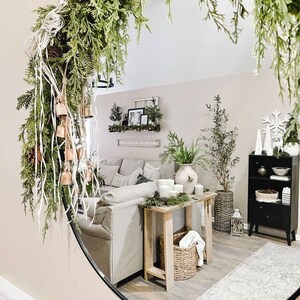 Peut inclure: Un miroir rond avec un cadre noir refl&egrave;te l'int&eacute;rieur d'un salon. Une guirlande verte avec des cloches est accroch&eacute;e en haut du miroir. Le reflet montre un canap&eacute; blanc, une table en bois avec des bougies, un panier en osier et une biblioth&egrave;que noire avec des objets d&eacute;coratifs.