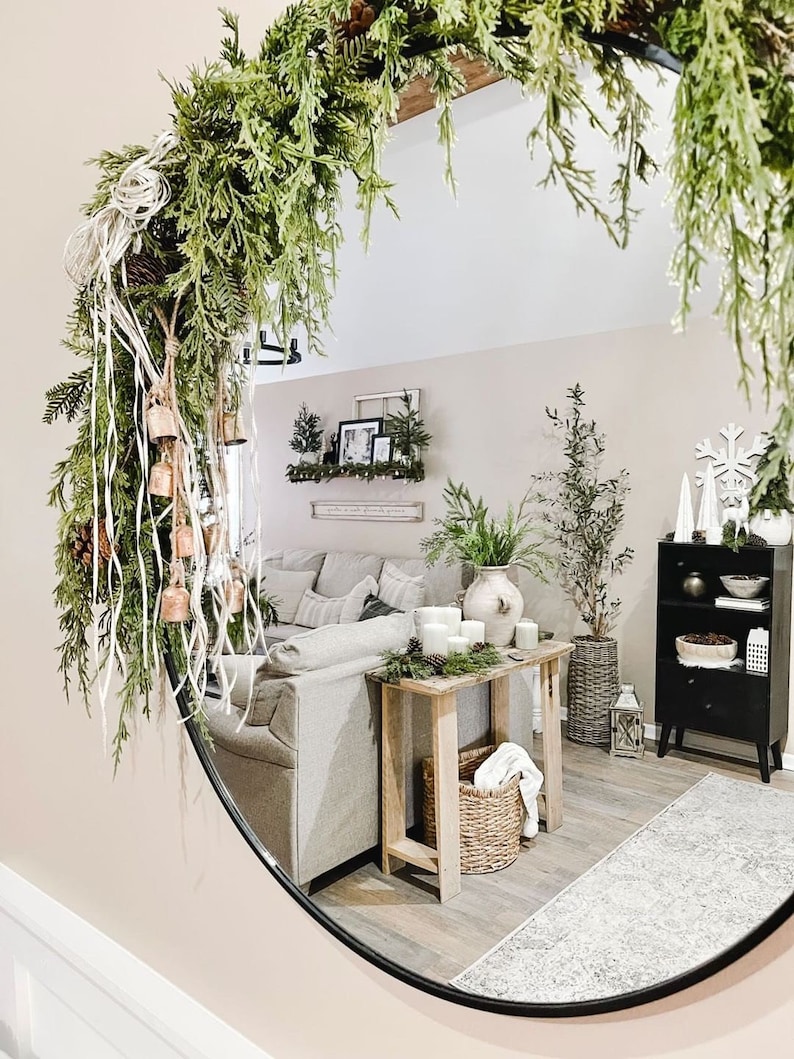 Peut inclure: Un miroir rond avec un cadre noir refl&egrave;te l'int&eacute;rieur d'un salon. Une guirlande verte avec des cloches est accroch&eacute;e en haut du miroir. Le reflet montre un canap&eacute; blanc, une table en bois avec des bougies, un panier en osier et une biblioth&egrave;que noire avec des objets d&eacute;coratifs.