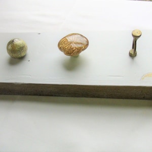 May include: A white wooden shelf with three decorative knobs. The knobs are a gold sphere, a brown glass knob with a floral design, and a gold hook.