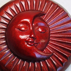 Puede incluir: Una talla de madera roja del sol y la luna con los ojos cerrados y bocas sonrientes. El sol tiene rayos alrededor.