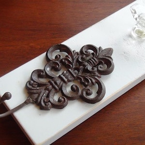 May include: A white wooden board with a decorative metal hook and a clear glass knob. The hook is shaped like a fleur-de-lis and has a brown finish.