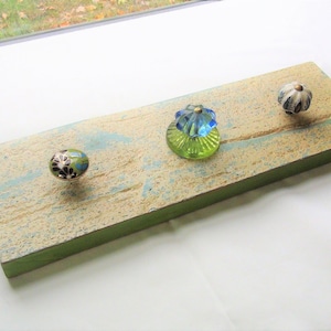 May include: A rustic wooden shelf with a green painted edge, featuring three decorative knobs. The knobs are made of glass and feature floral designs in shades of blue, green, and white.