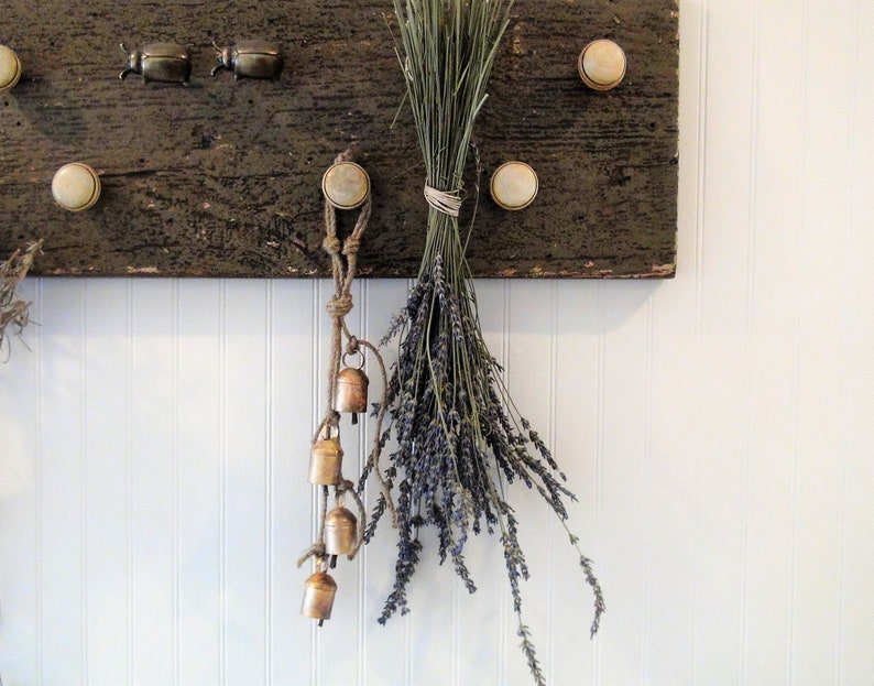 Peut inclure: Une planche de bois rustique avec des boutons en c&eacute;ramique blanche est accroch&eacute;e &agrave; un mur blanc. Un bouquet de lavande s&eacute;ch&eacute;e est suspendu &agrave; un bouton, et une cha&icirc;ne de petites cloches en laiton est suspendue &agrave; un autre bouton. Deux petits scarab&eacute;es en laiton sont fix&eacute;s &agrave; la planche.