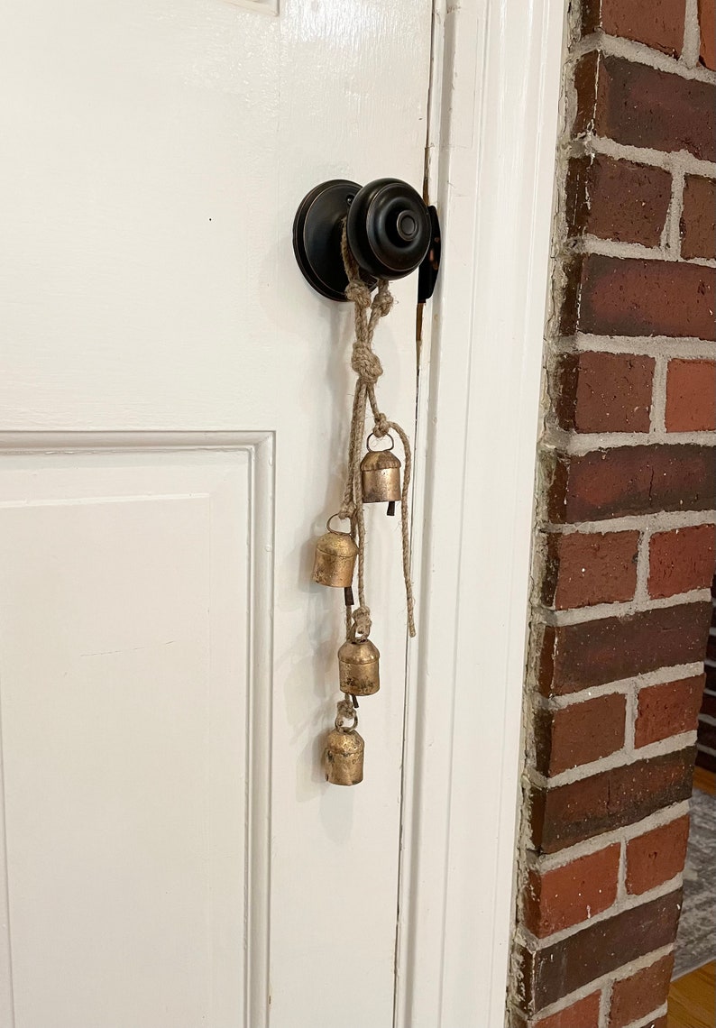Peut inclure: Une porte avec une poign&eacute;e de porte noire et une cha&icirc;ne de cloches dor&eacute;es qui pendent. Les cloches sont attach&eacute;es ensemble avec de la ficelle et pendent du bouton de porte.