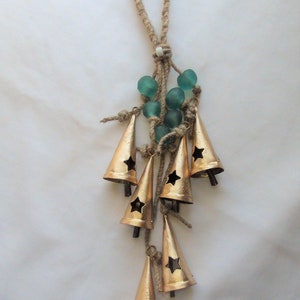 May include: A gold-colored metal wind chime with a star-shaped cutout on each bell. The chime is made of seven bells and is suspended from a length of twine with a loop at the top. The twine is decorated with teal-colored glass beads.