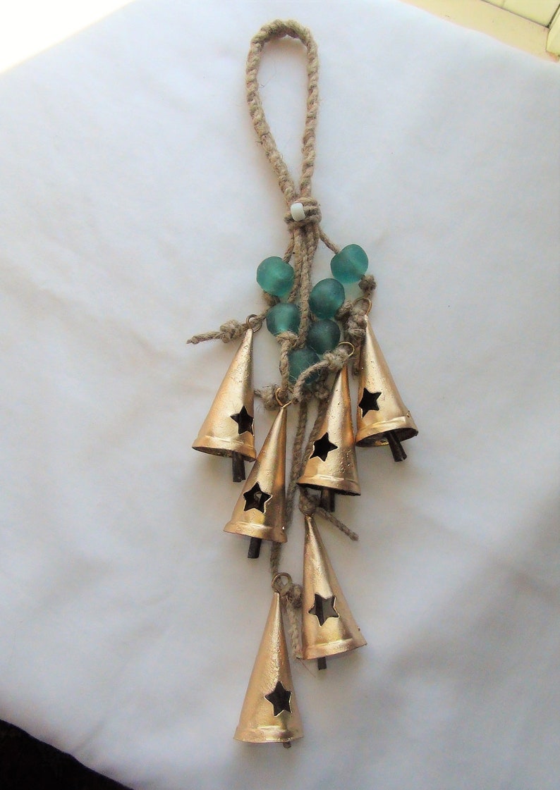 May include: A gold-colored metal wind chime with a star-shaped cutout on each bell. The chime is made of seven bells and is suspended from a length of twine with a loop at the top. The twine is decorated with teal-colored glass beads.