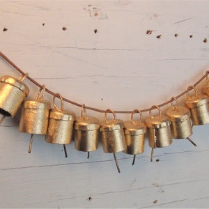 Op de afbeelding: Een rij van tien gouden belletjes die aan een bruin koord hangen. De belletjes zijn gelijkmatig verdeeld en hebben een kleine, puntige punt aan de onderkant.