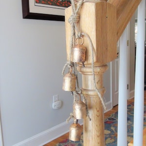 Peut inclure: Un poteau de contremarche en bois avec une corde et une s&eacute;rie de petites cloches dor&eacute;es qui pendent. Les cloches sont attach&eacute;es &agrave; la corde avec des n&oelig;uds. Le poteau fait partie d'un escalier avec une rampe blanche et un tapis &agrave; motifs au sol.