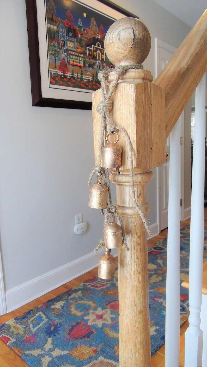 Peut inclure: Un poteau de contremarche en bois avec une corde et une s&eacute;rie de petites cloches dor&eacute;es qui pendent. Les cloches sont attach&eacute;es &agrave; la corde avec des n&oelig;uds. Le poteau fait partie d'un escalier avec une rampe blanche et un tapis &agrave; motifs au sol.