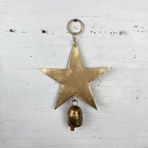 Op de afbeelding: Gouden metalen ster met een bel die eraan hangt. De ster hangt aan een ring op een witte houten achtergrond.
