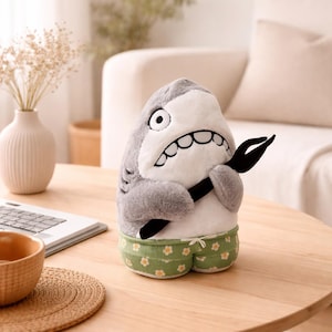 Puede incluir: Un juguete de peluche de tiburón con cuerpo gris, vientre blanco y boca muy abierta sosteniendo una herramienta negra. El tiburón lleva pantalones cortos verdes con estampado floral. El juguete está sobre una mesa de madera.
