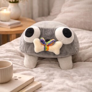 May include: A gray plush toy with large white eyes and black pupils, resembling a cartoon character. It has a cream-colored bone-shaped detail with a rainbow ribbon. The toy is on a bed with a neutral-colored comforter.
