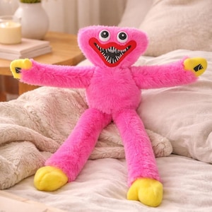 Puede incluir: Un peluche rosa con una cara sonriente, ojos blancos grandes y una boca roja ancha llena de dientes negros. El juguete tiene brazos y piernas largos, con manos y pies amarillos. Está colocado sobre una cama con una manta de color neutro.