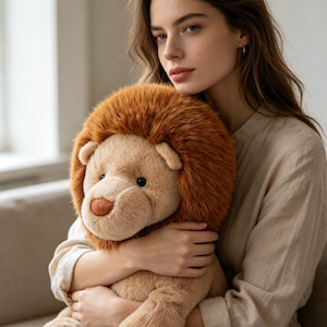 May include: A plush lion toy with a brown mane and a tan body. The stuffed animal has a friendly expression with a soft, cuddly appearance. The toy is being held by a person.