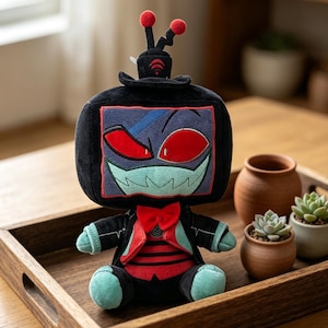 Op de afbeelding: Een pluche speelgoed in de vorm van een televisie met een dreigend gezicht, rode ogen en een vlinderdas. Het speelgoed is zwart met rode en blauwe accenten en staat op een houten dienblad. Kleine plantenpotten zijn zichtbaar op de achtergrond.