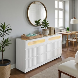 May include: A white modern dresser with a ribbed design and two sets of drawers. The dresser has a glass top and a warm light feature. A round gold mirror hangs above the dresser, and a woven basket with blue decorative items sits on top.