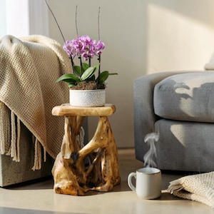 May include: A small, rustic wooden stool supports a white ceramic pot filled with a vibrant purple orchid. A steaming white mug sits nearby, suggesting a cozy, relaxing atmosphere. A beige throw blanket and a gray sofa complete the scene.