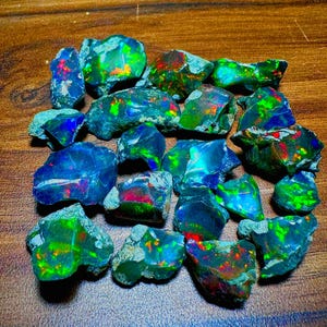 Op de afbeelding: Een verzameling ruwe, donkergekleurde opaalstenen met levendige flitsen van iriserend groen, blauw en rood kleurenspel. Elke steen heeft een unieke, onregelmatige vorm met een gepolijst oppervlak dat licht reflecteert en diepe tinten laat zien.