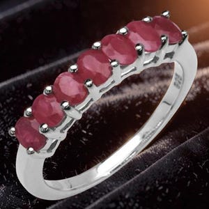 May include: A silver ring featuring a row of seven oval-cut red rubies set in a prong setting. The band is polished silver and includes a 925 hallmark stamp on the inner side, indicating sterling silver composition for this jewelry piece.