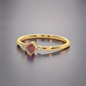 May include: A gold ring featuring a central round-cut red gemstone flanked by two smaller clear round-cut stones. The band is smooth and polished, with the stones set in a delicate arrangement that highlights the contrast between the red and clear gems.