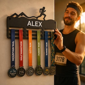 Op de afbeelding: Een zwarte metalen medaillehouder met de naam ALEX in witte letters, een silhouet van een hardloper en bergtoppen. Zes kleurrijke linten hangen aan het rek, elk bevestigd aan een ronde medaille met racenamen zoals Mountain Trail, Half Marathon, City Run, Forest Run, Sunset Run en Marathon 42K.
