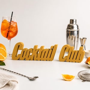 Peut inclure: Une enseigne tridimensionnelle jaune moutarde avec le texte Cocktail Club dans une police grasse sans empattement. L'enseigne est présentée avec un verre de cocktail orange avec une paille noire, un shaker en argent, un doseur et une passoire.