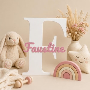 Pode incluir: Uma letra F grande de madeira branca com o nome Faustine em letras cursivas cor-de-rosa fixado na frente. Ao lado, há um coelho de peluche bege, uma almofada em forma de estrela com cara de sono, um arco-íris de madeira e um chocalho.