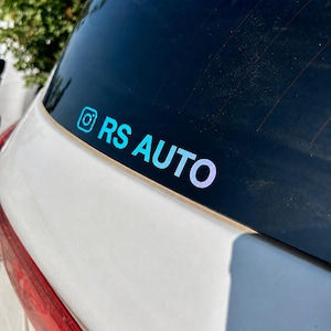 Può includere: Un adesivo olografico applicato sul lunotto posteriore di un veicolo bianco. L'adesivo presenta l'icona del logo Instagram seguita dal testo RS AUTO in un carattere sans-serif in grassetto. Le lettere mostrano un effetto sfumato cangiante.