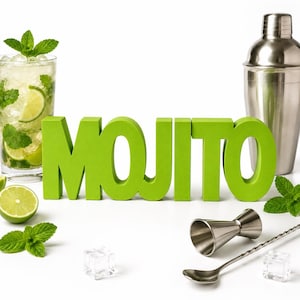 Könnte beinhalten: Ein hellgrünes Schild mit dem Wort MOJITO in Blockbuchstaben, platziert neben einem Glas mit einem Limetten-Minz-Cocktail, einem Cocktailshaker aus Edelstahl, einem Messbecher, einem langen Rührlöffel und verstreuten Eiswürfeln.