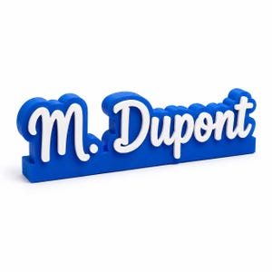Peut inclure: Une enseigne en trois dimensions affichant le nom M. Dupont en lettres cursives blanches sur une base bleue unie. Les lettres présentent un aspect en relief et superposé avec une finition lisse, créant un contraste net entre le texte blanc et le fond bleu.