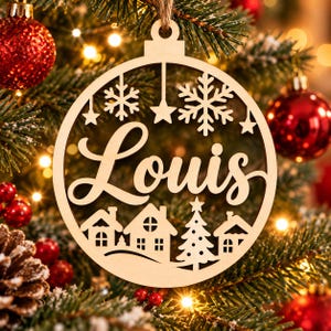 Peut inclure: Une décoration de Noël circulaire en bois portant le prénom Louis en écriture cursive. Le motif comprend des découpes de flocons de neige, d'étoiles, de petites maisons et d'un sapin, suspendue par une boucle en ficelle.