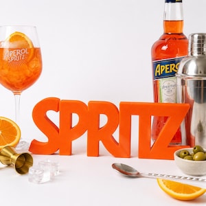Peut inclure: Une enseigne 3D orange vif formant le mot SPRITZ, placée à côté d'un verre d'Aperol Spritz, d'une bouteille d'Aperol, d'un shaker en métal, d'un doseur doré, d'une cuillère argentée, de deux glaçons, d'un bol d'olives vertes et de tranches d'orange.