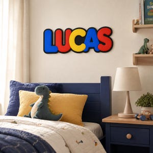 Könnte beinhalten: Ein Wandschild mit dem Namen LUCAS in fetten, abgerundeten, dreidimensionalen Buchstaben. Die Buchstaben sind in einer Abfolge von Blau, Rot, Gelb, Blau und Rot gefärbt und jeweils mit einem dicken schwarzen Rand an der Wand versehen.