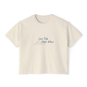 May include: Cream-colored t-shirt with the text "Low Tide High Vibes" in navy blue. The design includes a wave graphic. The shirt has a crew neck and short sleeves, perfect for casual wear.