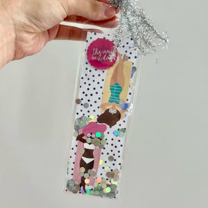 May include: A clear plastic bag filled with confetti and two illustrations of people sunbathing. The bag has a white background with black polka dots. A pink circle with the text "the very best day" is at the top. Silver tinsel is attached to the top.